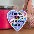 Heart-shaped sticker with text next to a pink candle and vase with flowers on a wooden surface.