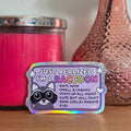 Colorful sticker with text on a wooden surface next to a pink candle and vase with flowers.