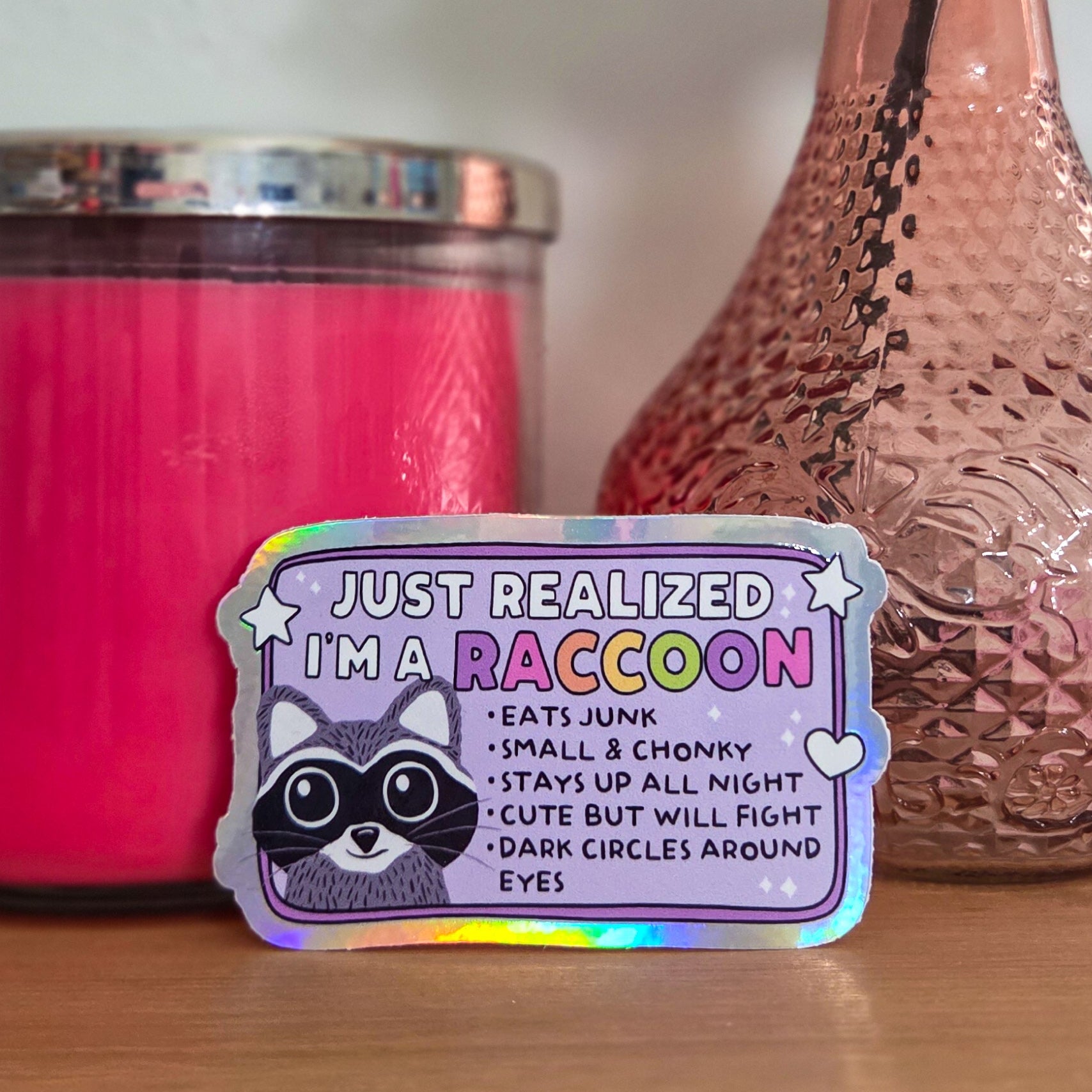 Colorful sticker with text on a wooden surface next to a pink candle and vase with flowers.