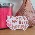 Clear sticker with 'I'm trying my best honey' text on a wooden surface with a pink candle and vase in the background.
