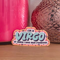 A decorative STICKER with the text 'I'M A VIRGO OF COURSE I HAVE HIGH STANDARDS' in light blue and black lettering, styled on a wooden surface with a pink candle and a brown textured vase in the background.